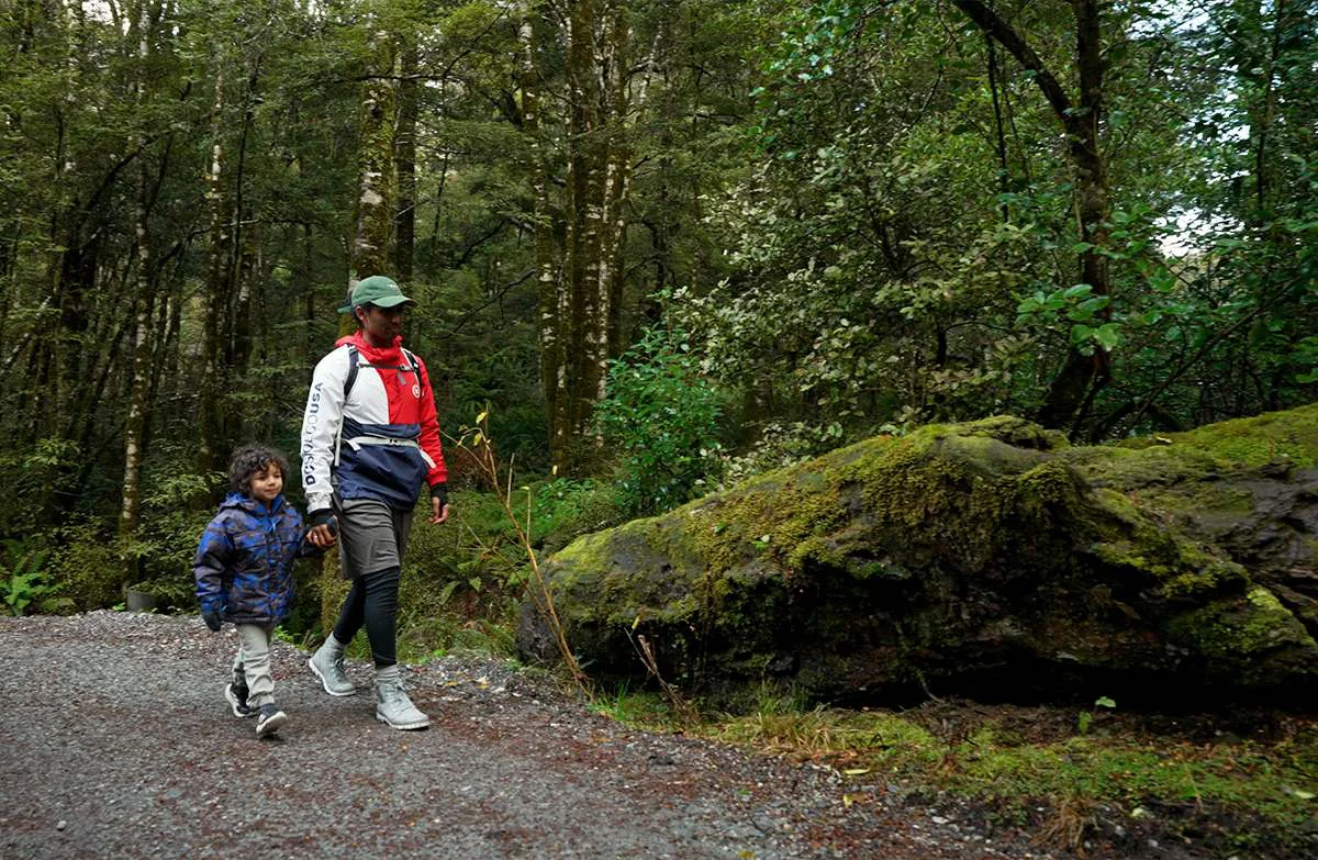 An adult and child walk hand in hand along a gravel path past a large mossy rock.
