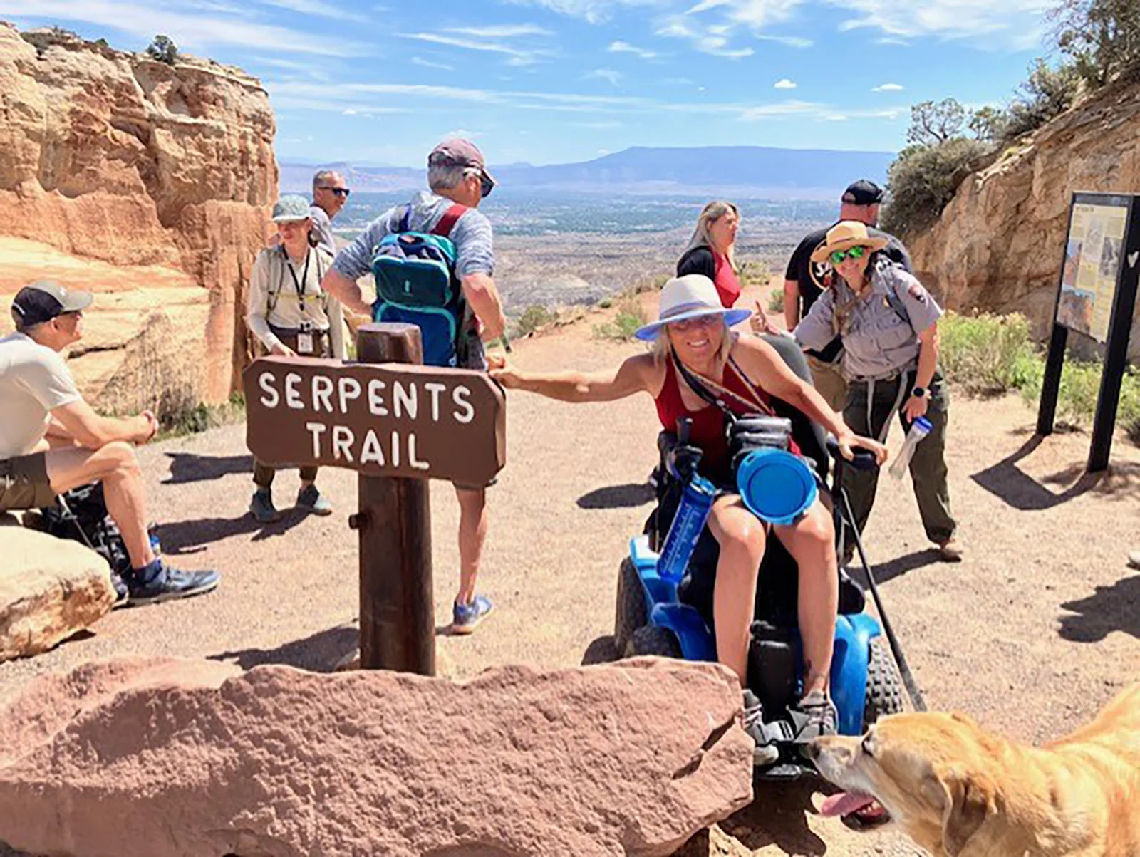 woman using wheelchair and service animal points to Serpents Trail sign with uniformed ranger behind