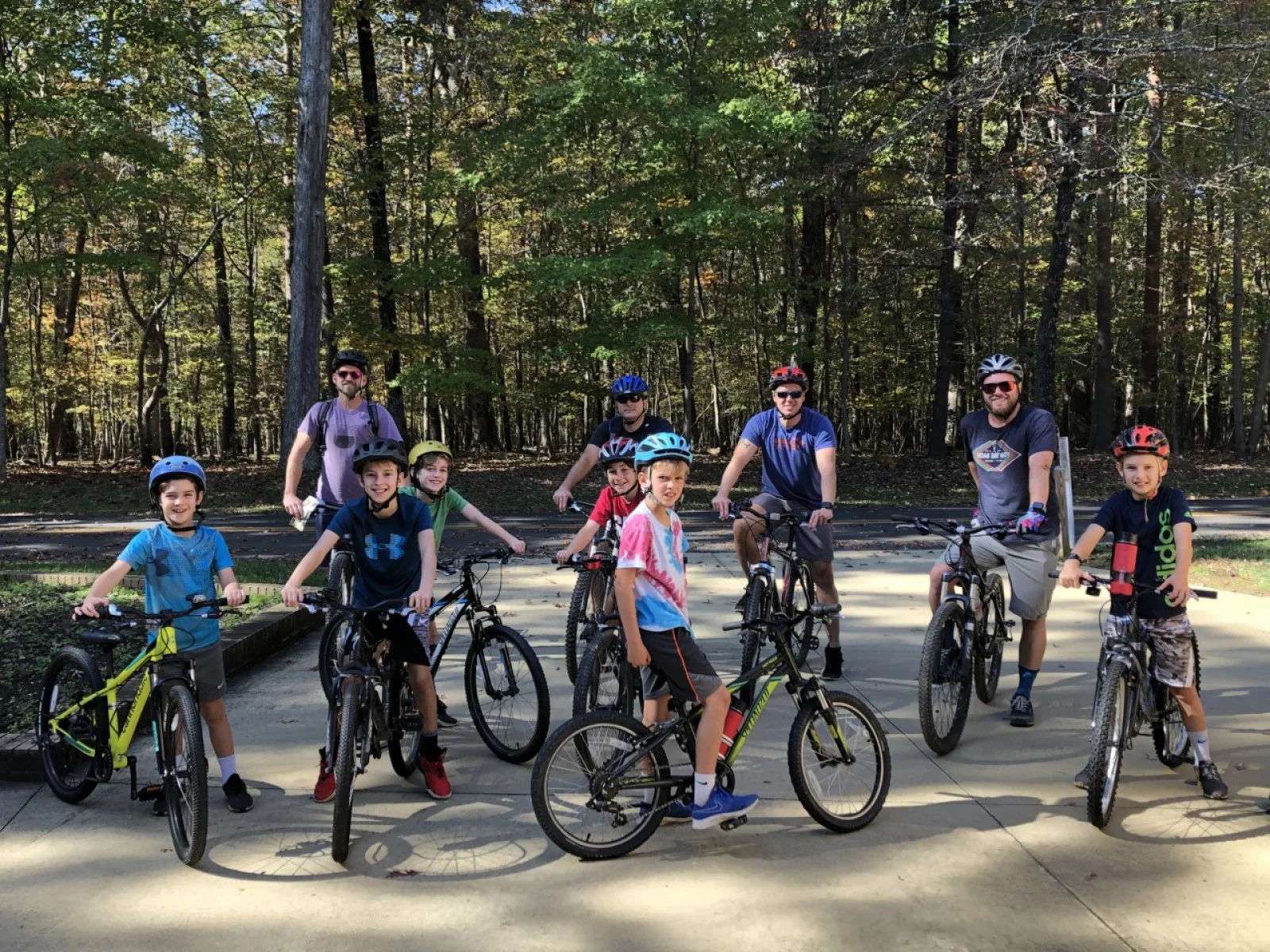 Ten helmeted cyclists pose with their bicycles on a paved road beside woodland.
