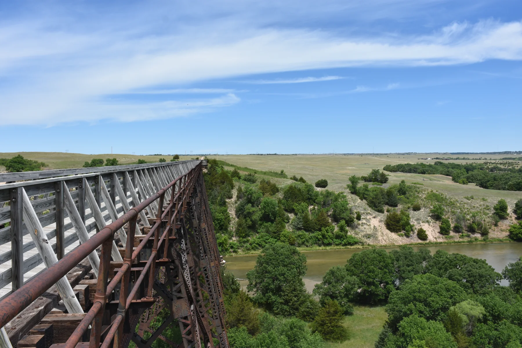 A metal bridge with wood walkway extends across a green landscape with a river below.