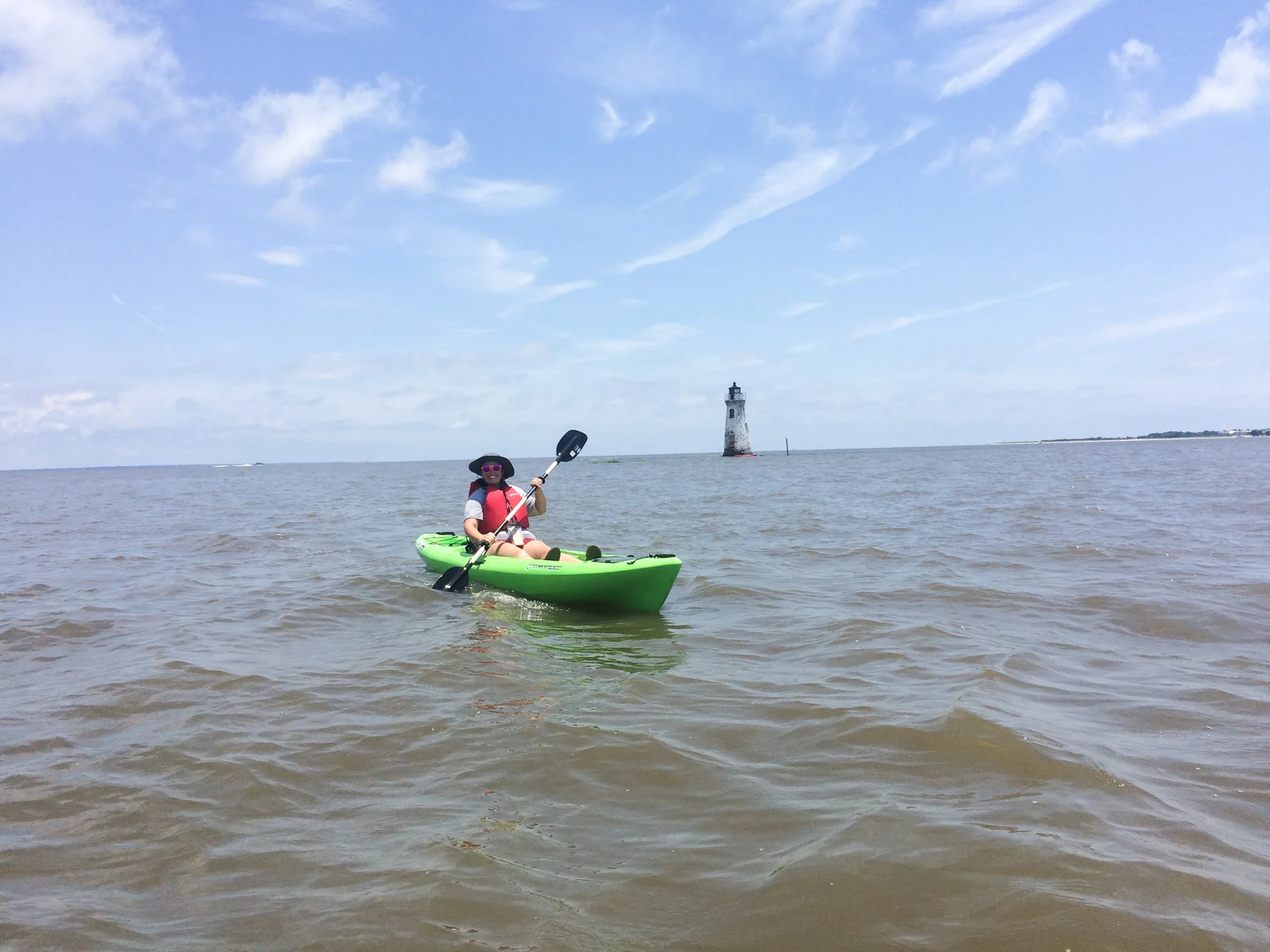 A woman in a green kayak with a lighthouse distantly behind.