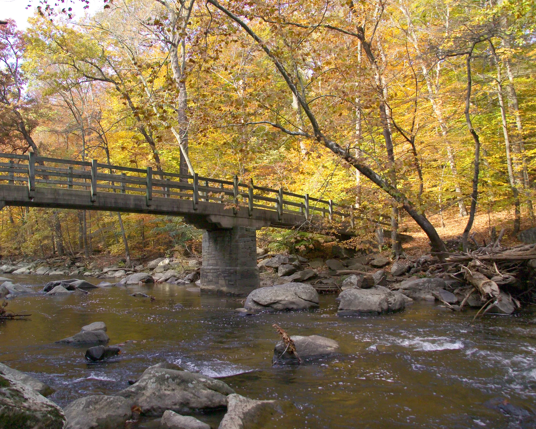 A wood and concrete bridge crosses a rocky creek with yellow and green trees on the far bank