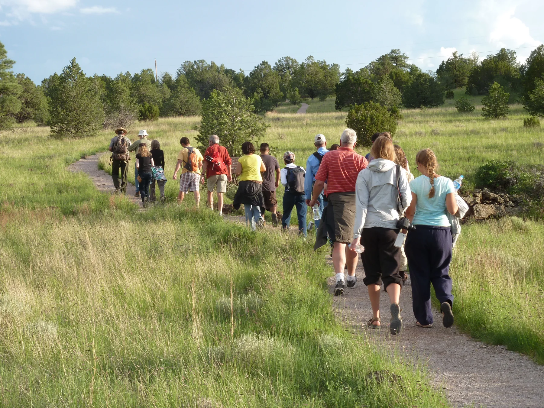 A group of hikers heads down a gravel trail through grass with trees in the distance